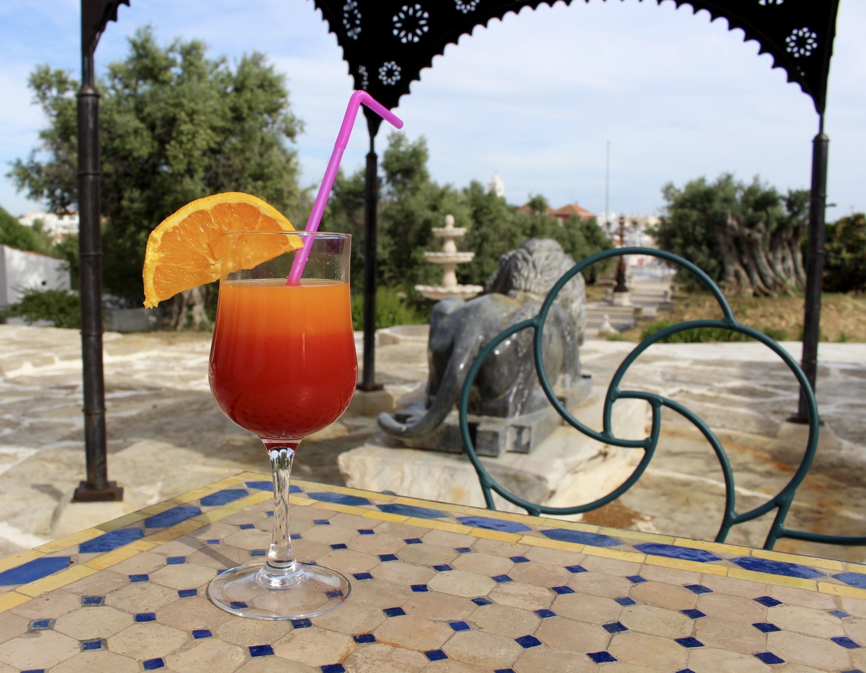 A glass of red beverage with a straw and an orange slice on a tiled table outdoors.