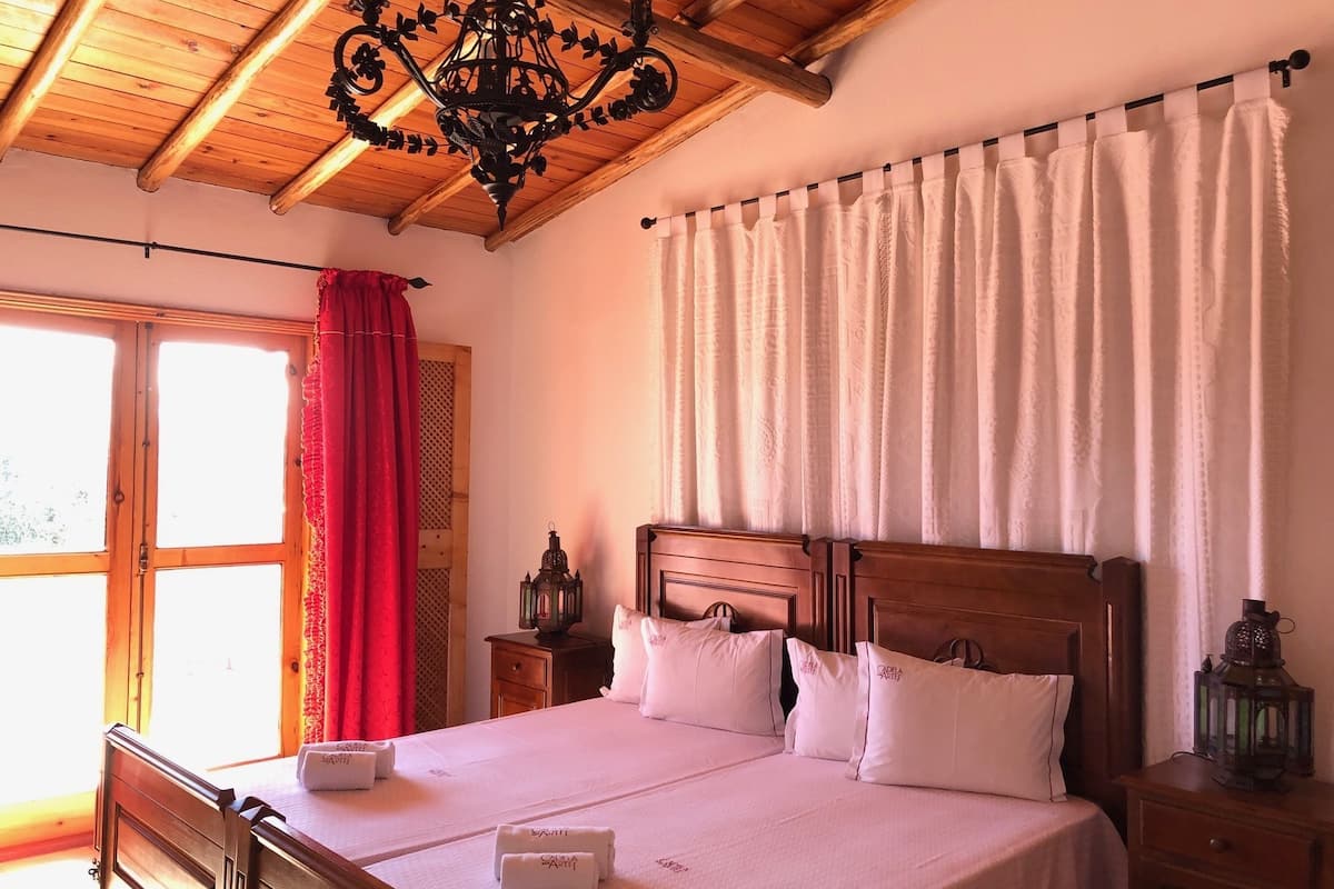 Ein Schlafzimmer mit Holzbalkendecke, einer Chandelieber, einem Bett mit weißen Leinen und einem Fenster mit rotem Vorhang.