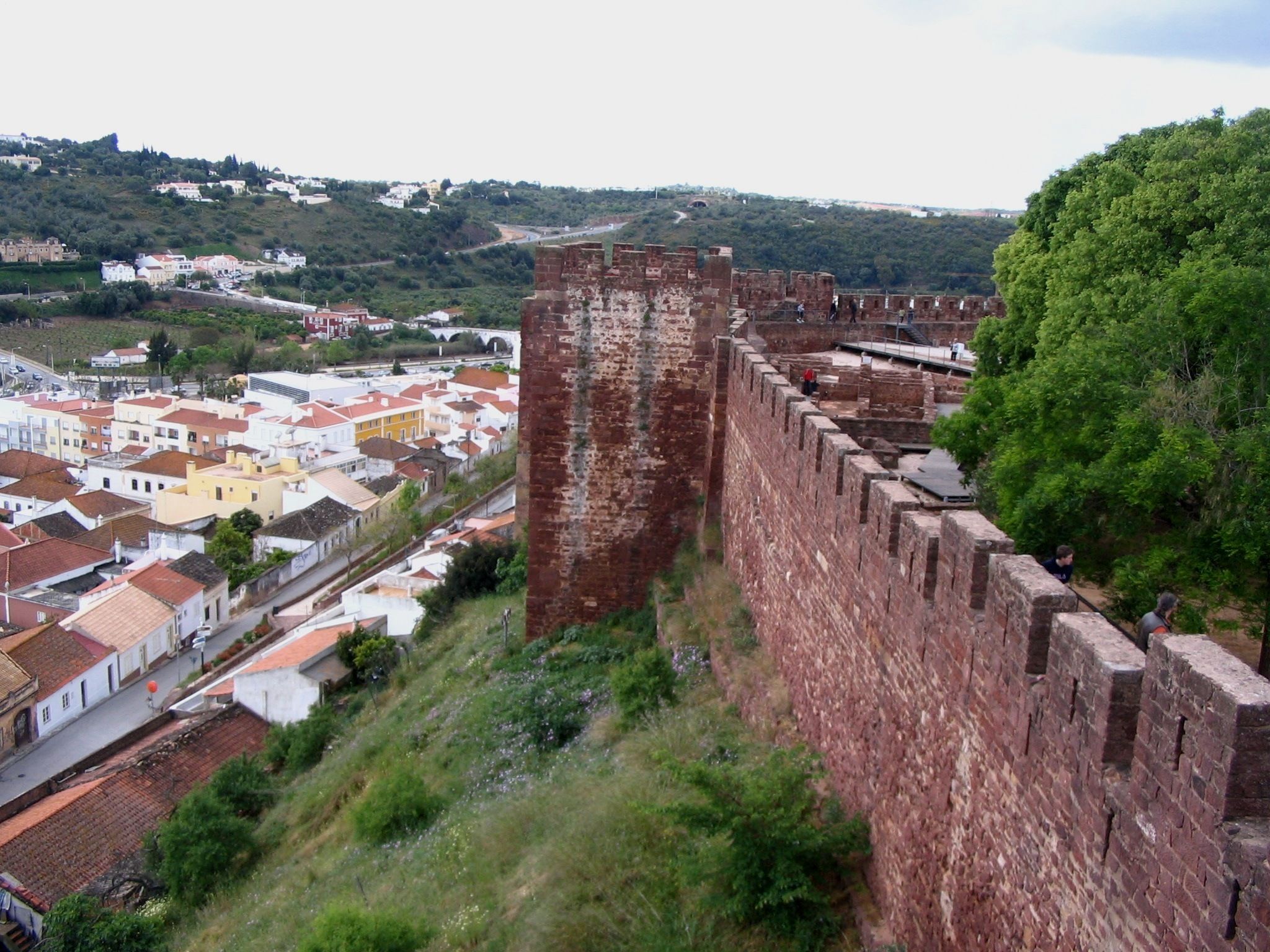 A historic stone wall with a fortified structure, overlooking a town with colorful buildings and a hilly landscape in the background.