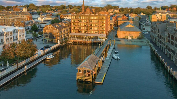 A riverside town with a prominent wooden pier, a large building with multiple windows, and a church spire in the background.
