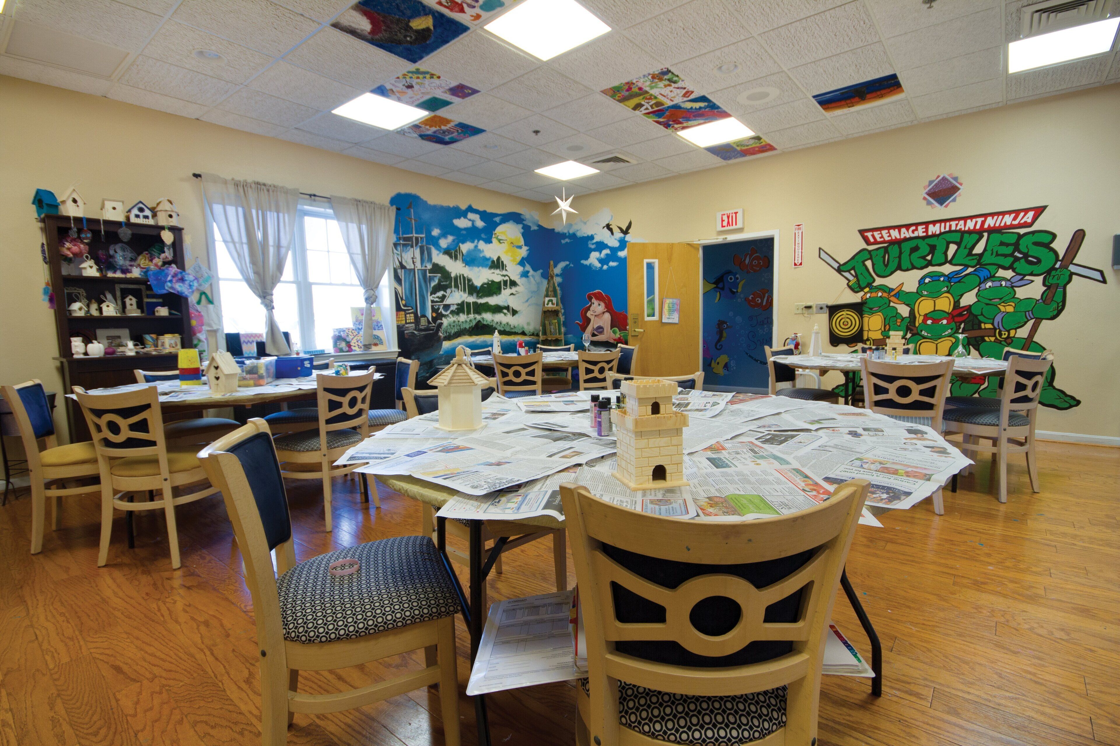 A classroom with wooden chairs and tables, educational posters, and a mural on the wall.