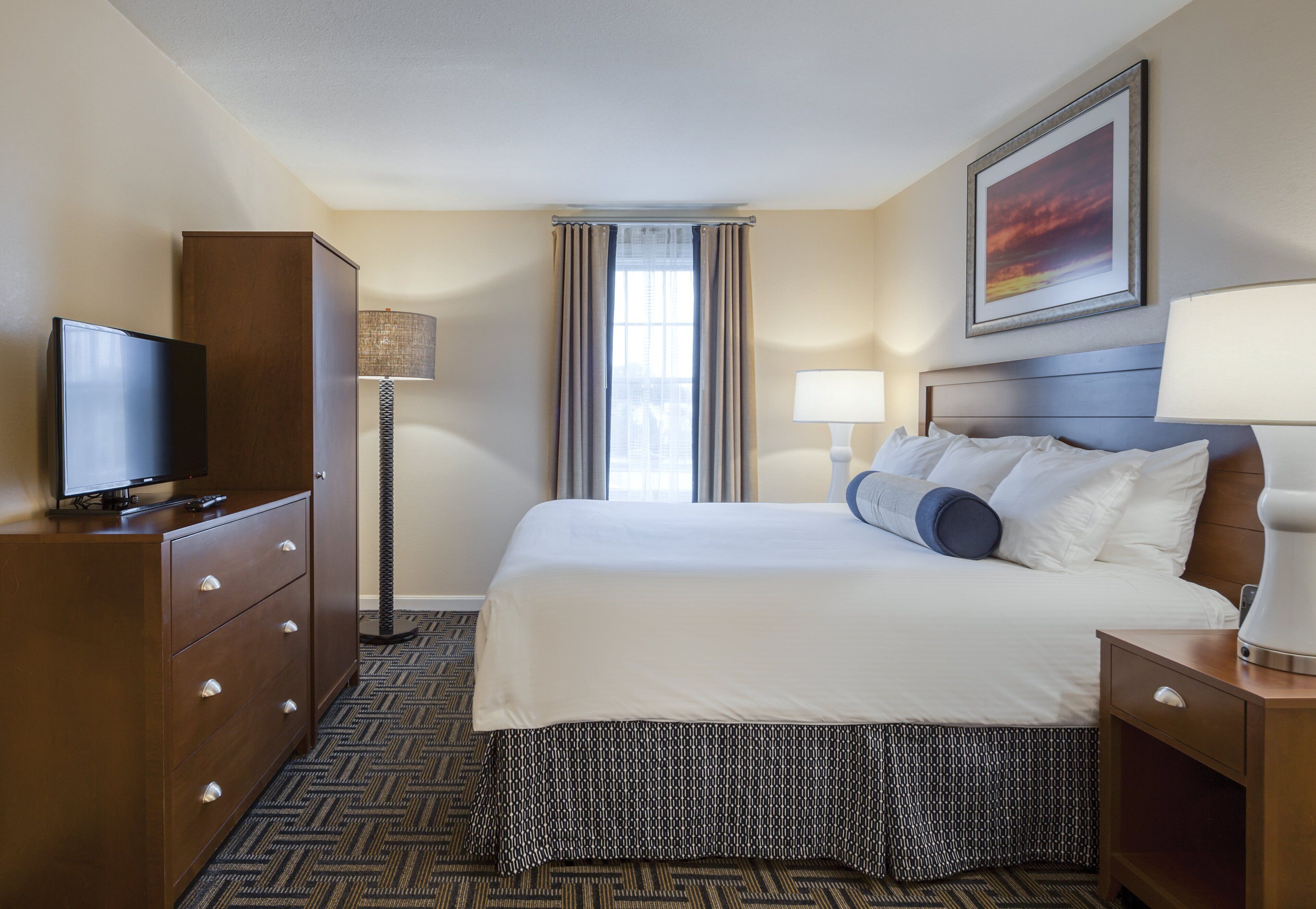 A hotel room with a bed, a TV on a dresser, a lamp, and a painting on the wall.
