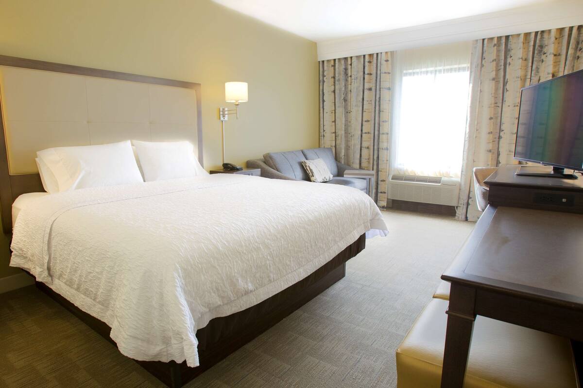 Een hotelkamer met een groot bed, een bureau met een computer, een stoel, een lamp en een raam met gordijnen.