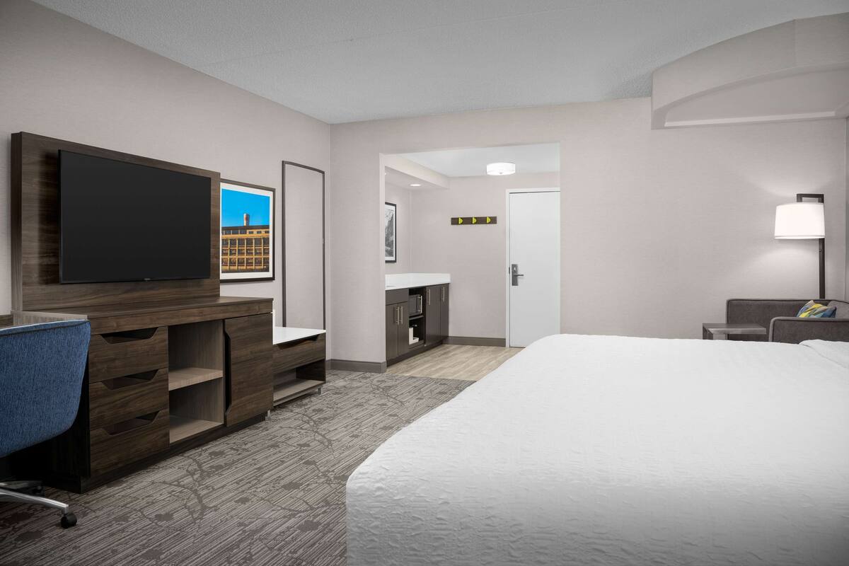 A hotel room with a large bed, a desk with a flat-screen TV, a chair, a desk lamp, and a view of a building through the window.