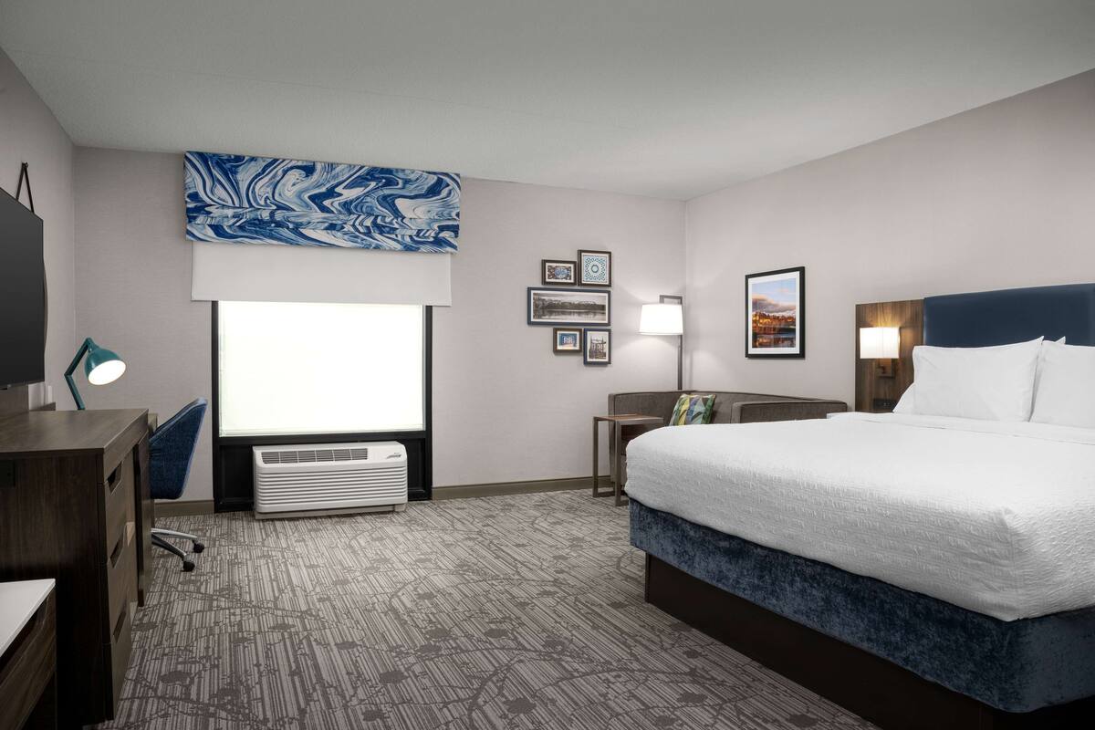A hotel room with a large bed, a desk, a chair, a flat-screen TV, a wall-mounted air conditioner, and framed pictures on the wall.
