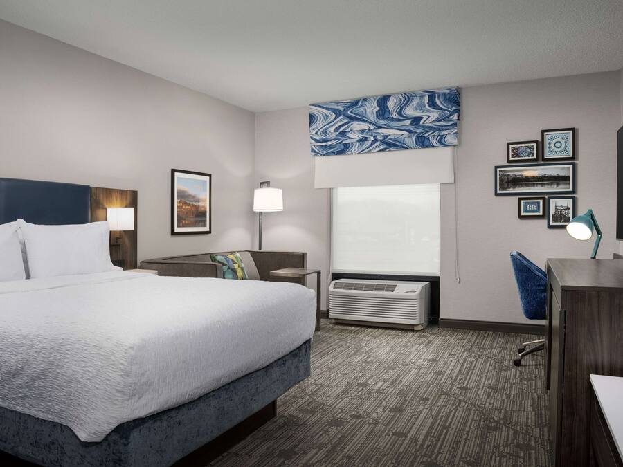 Ein Hotelzimmer mit einem Bett, einem Schreibtisch, einem Stuhl, einer Lampe, einer Wand-Klimaanlage und gerahmten Bildern.
