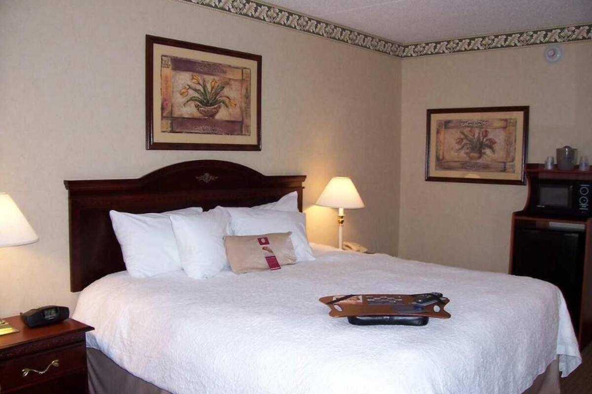 A neatly made bed with white linens, a wooden headboard, two bedside lamps, and a small table with a remote control.