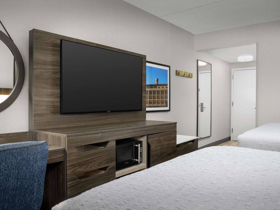 Habitación de hotel con televisor de pantalla plana montado en una consola de madera, microondas integrado en la consola, cama con ropa blanca y puerta que da a otra habitación.