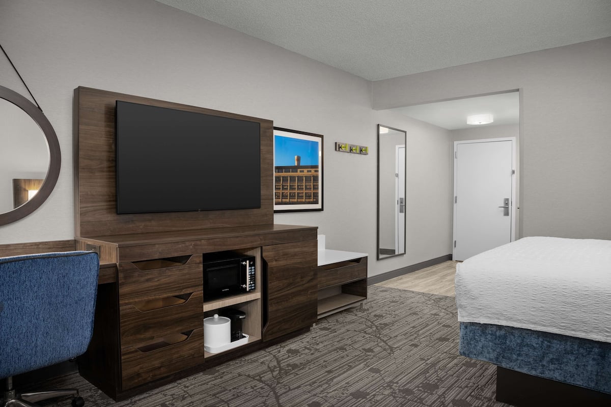 A hotel room with a flat-screen TV mounted on a wooden cabinet, a desk with a chair, a bed with a white comforter, and a door leading to another room.