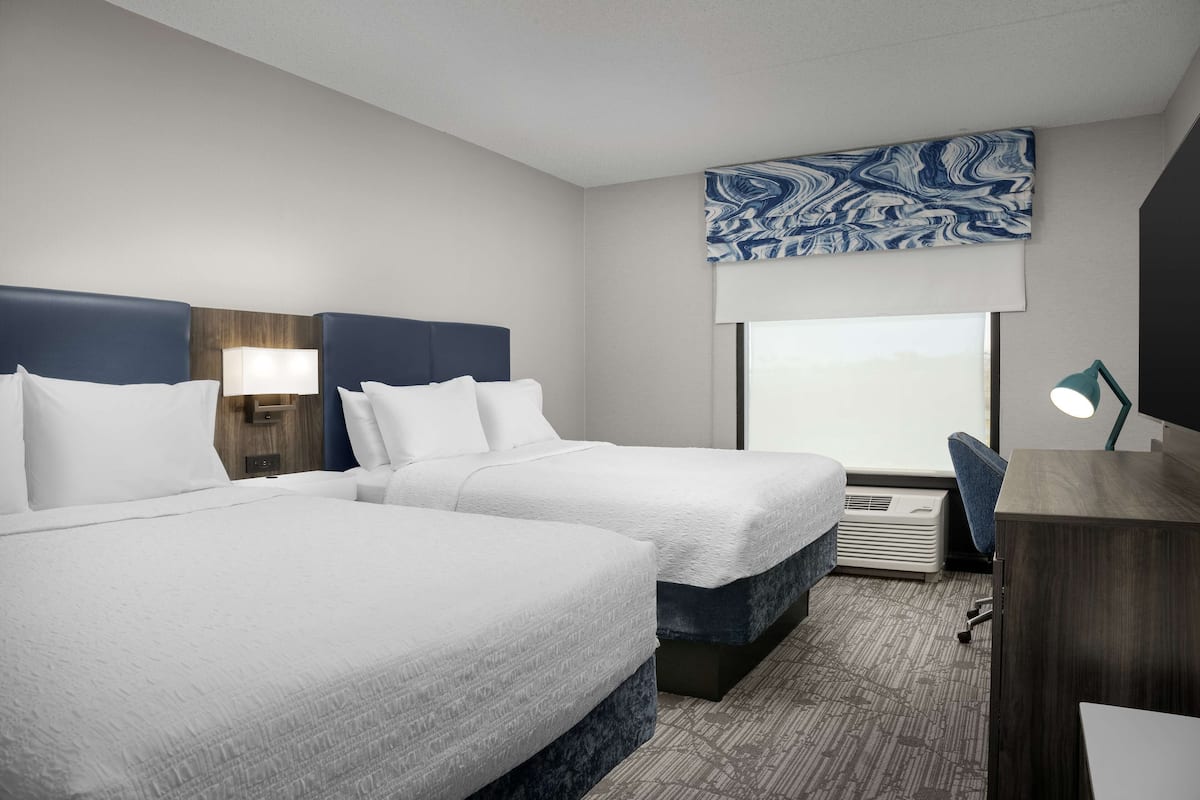 A hotel room with two beds, a desk, a chair, a large window, and a wall-mounted artwork.
