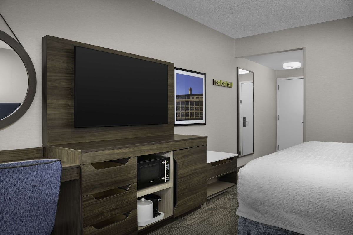 A hotel room with a large flat-screen TV mounted on a wooden cabinet, a bed with a white comforter, a small desk with a chair, and a framed picture on the wall.