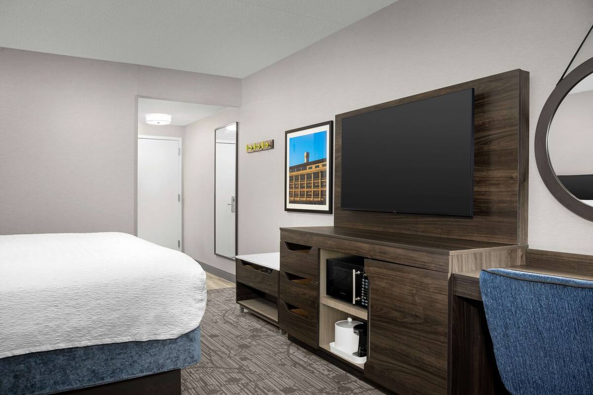 A hotel room with a bed, a TV, a mirror, and a small desk with drawers.