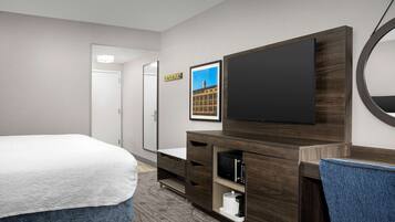 A hotel room with a bed, a TV, a mirror, and a small desk with drawers.
