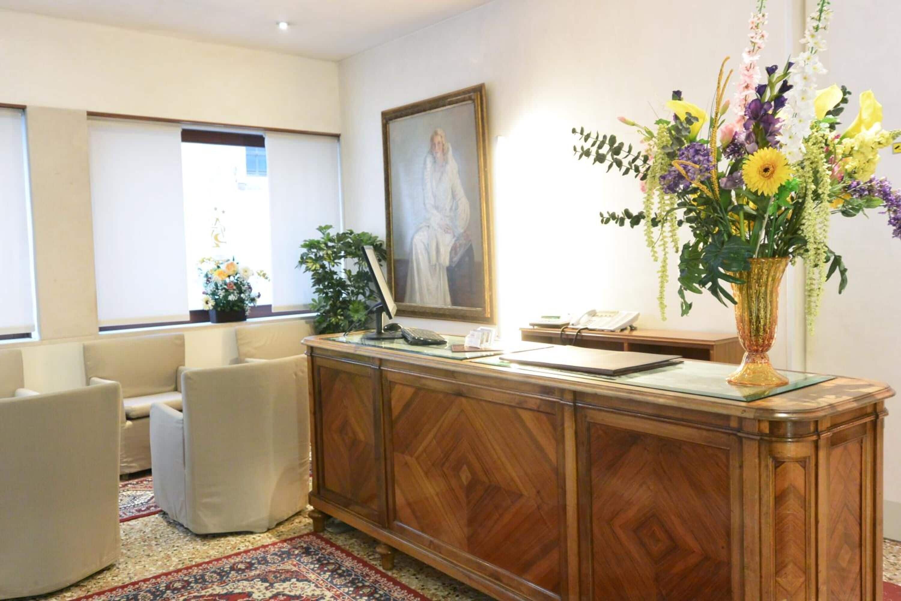 A reception area with a wooden desk, a framed portrait, a vase of flowers, and a patterned rug.