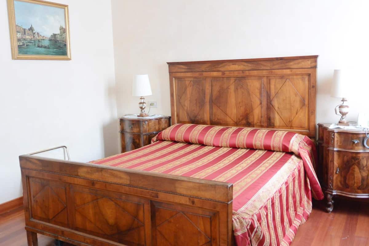 A wooden bed with a red and gold striped bedspread, a wooden nightstand with a lamp, and a wooden dresser with a mirror.