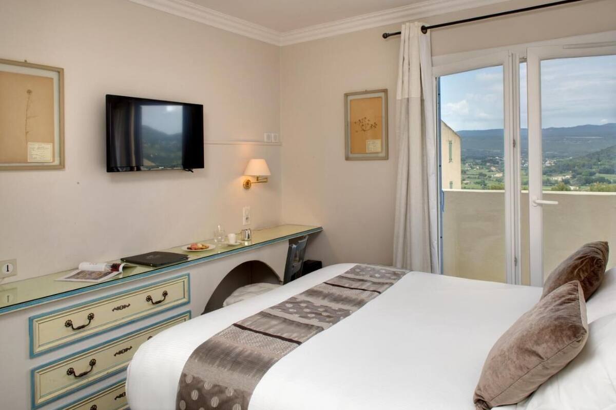 Een hotelkamer met een bed, een bureau, een televisie en een balkon met uitzicht.