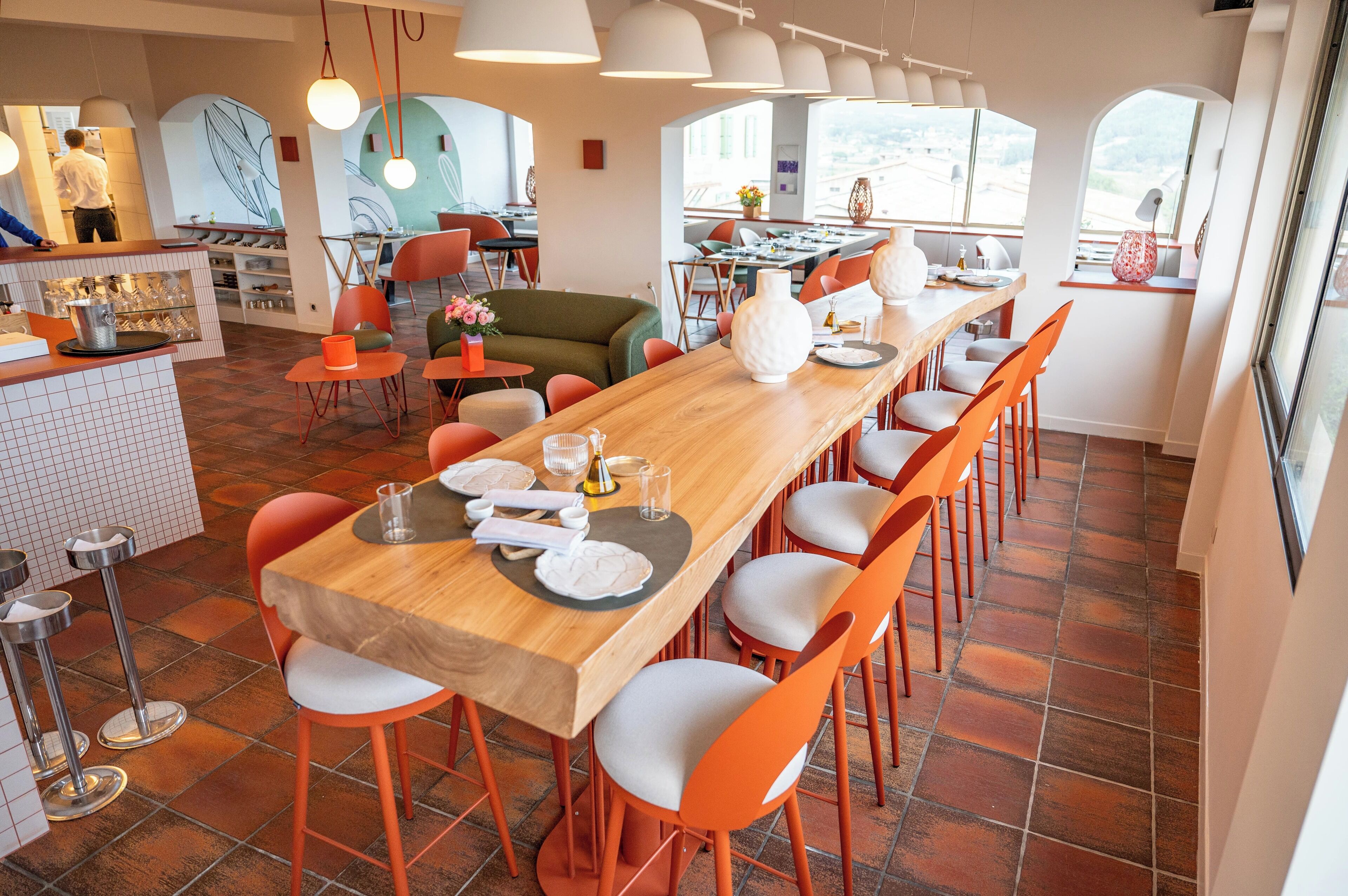 Un espace repas moderne avec une longue table en bois, des chaises orange et un coin bar avec des tabourets.