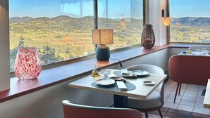 Un coin repas avec une table dressée pour deux personnes, un vase et une lampe. De grandes fenêtres offrent une vue sur la campagne.