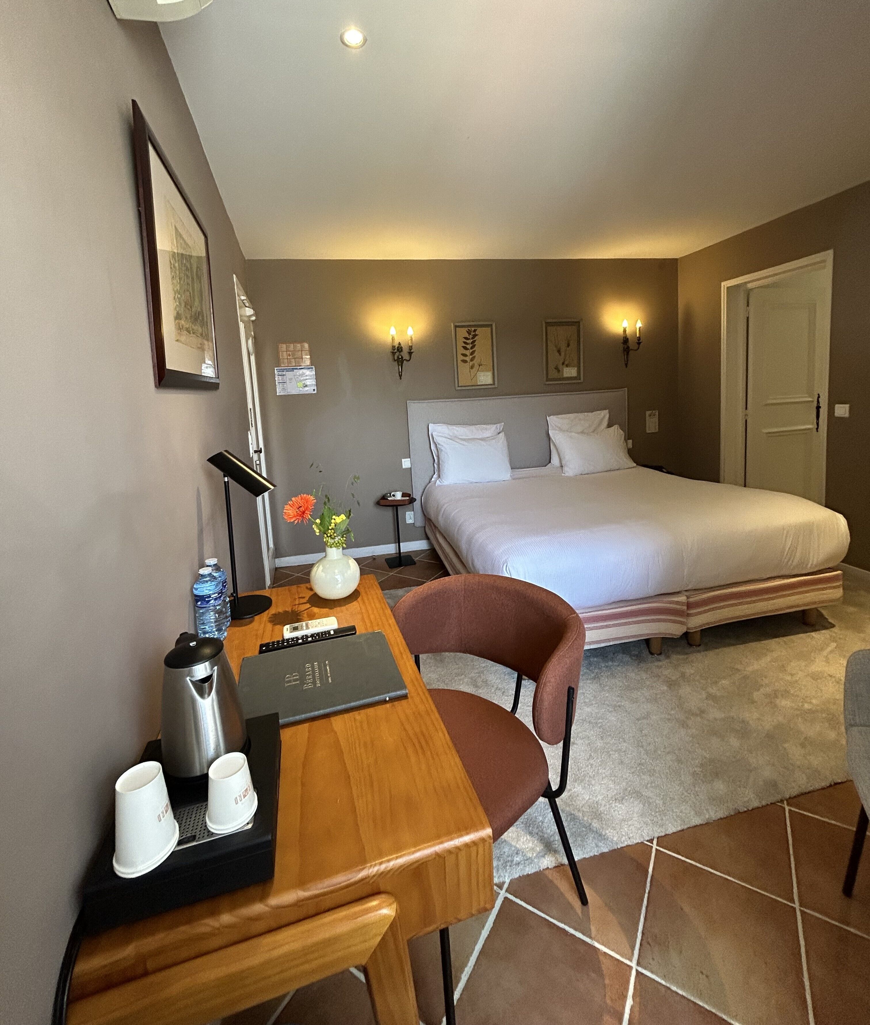 A hotel room with a bed, a desk with a laptop, a chair, a coffee maker, and a vase with flowers.