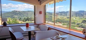 A dining area with a table and chairs, a lamp, and a view of a hilly landscape through large windows. - Maison Bérard – hôtel restaurant spa (La Cadiere-d'Azur)