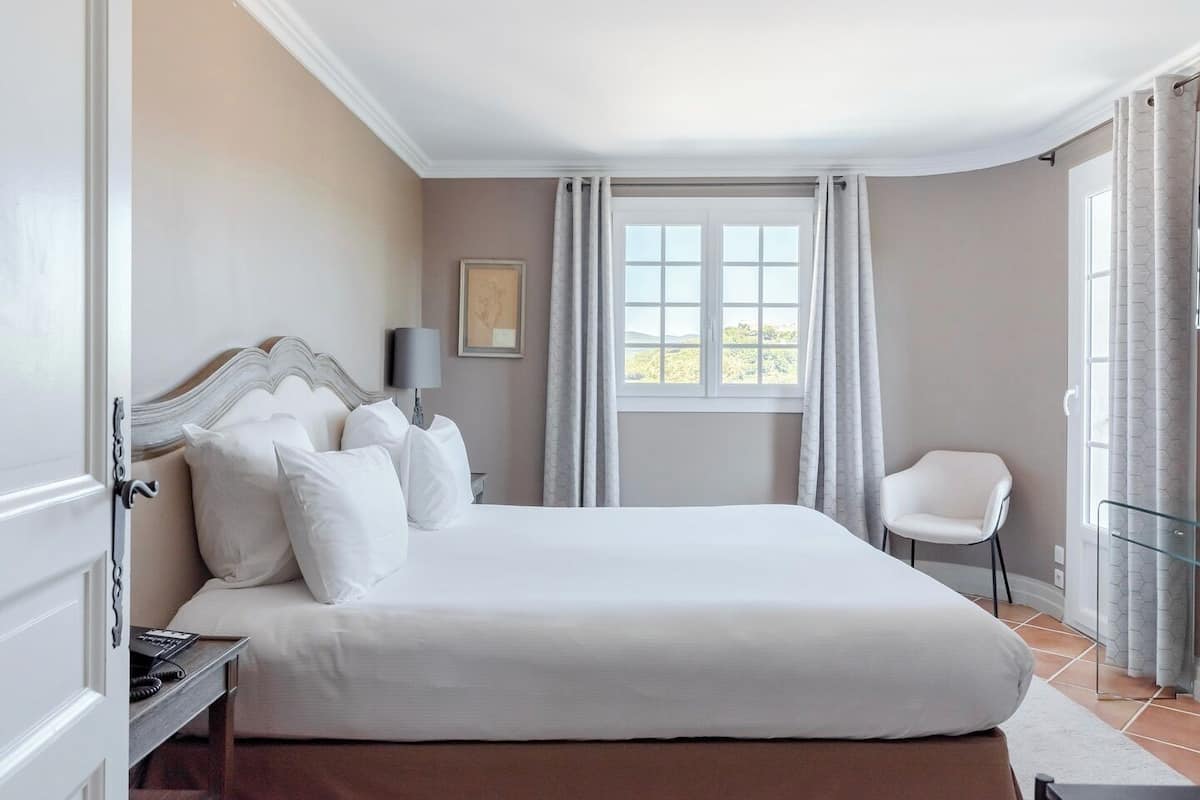 Een slaapkamer met een groot bed, een nachtkastje, een stoel, een raam met gordijnen en een ingelijst schilderij aan de muur.