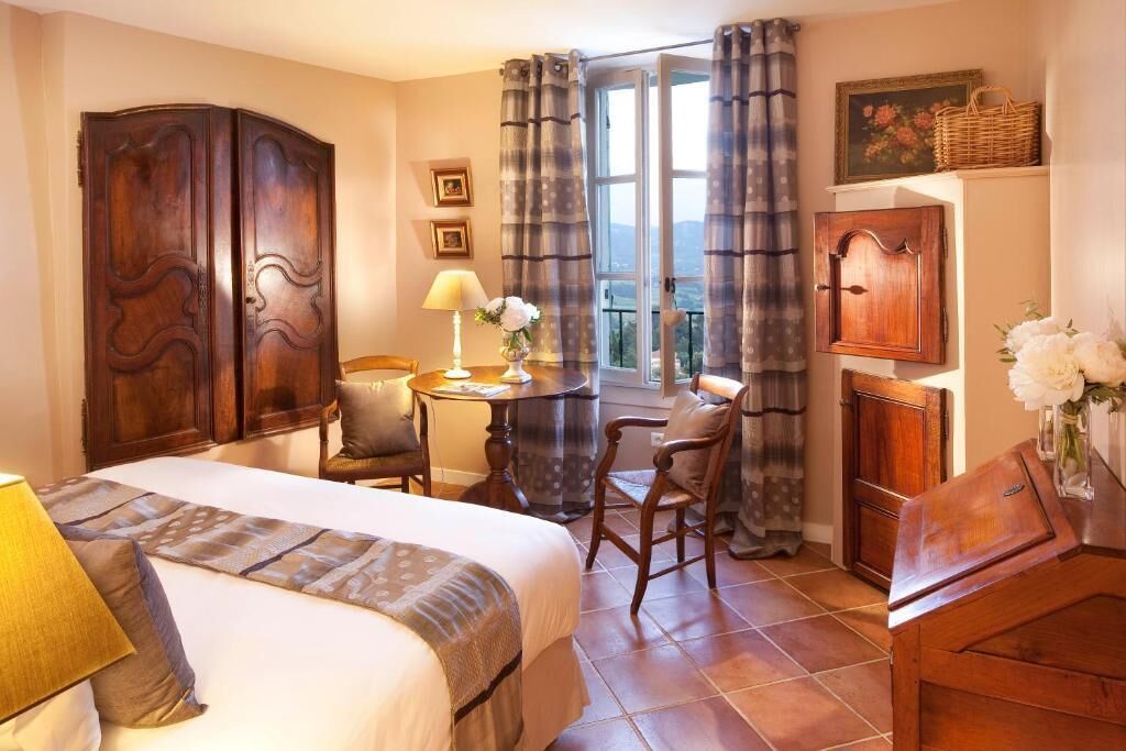 A bedroom with a bed, a desk, two chairs, a lamp, a window with curtains, and a view of the outdoors.