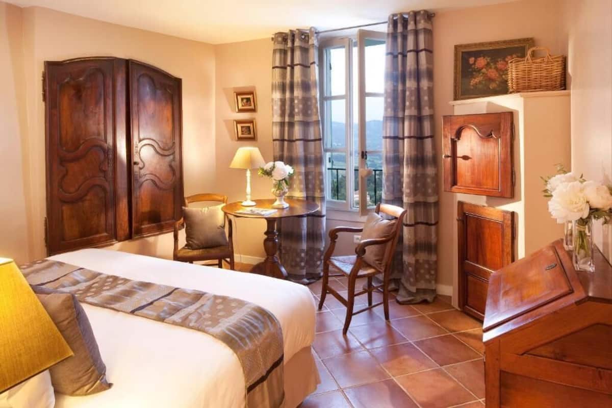 Een slaapkamer met een bed, een bureau, twee stoelen, een lamp, een raam met gordijnen en uitzicht op de omgeving.
