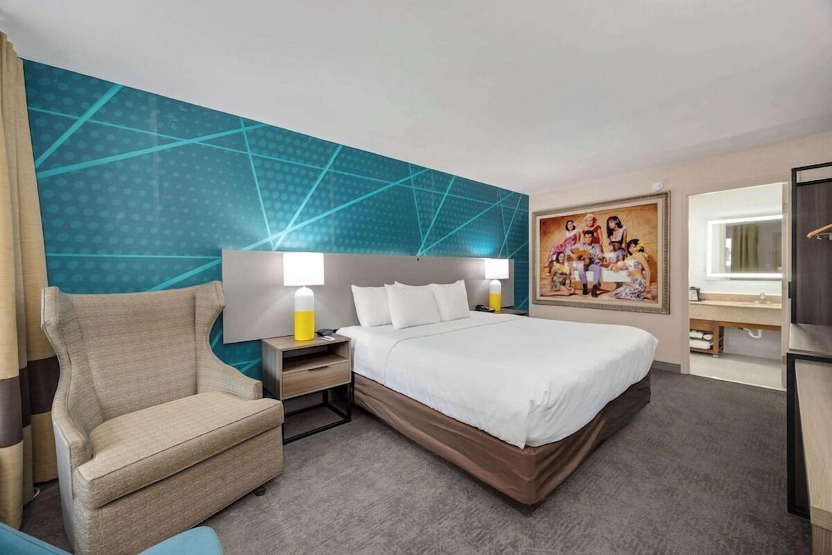 A hotel room with a large bed, a chair, a nightstand, a painting, and a window.