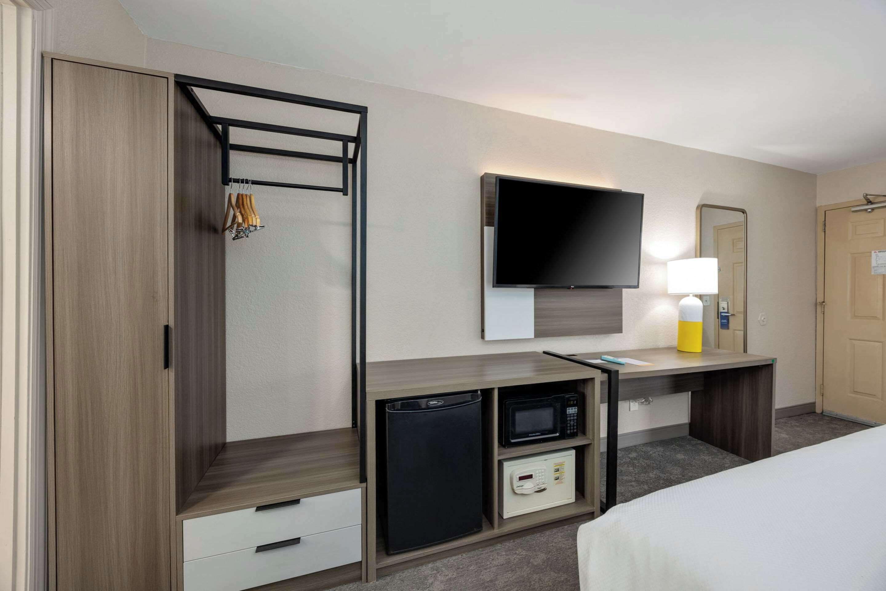 A hotel room with a bed, a TV mounted on the wall, a desk with a microwave, a mini-fridge, and a wardrobe with a hanging rack.