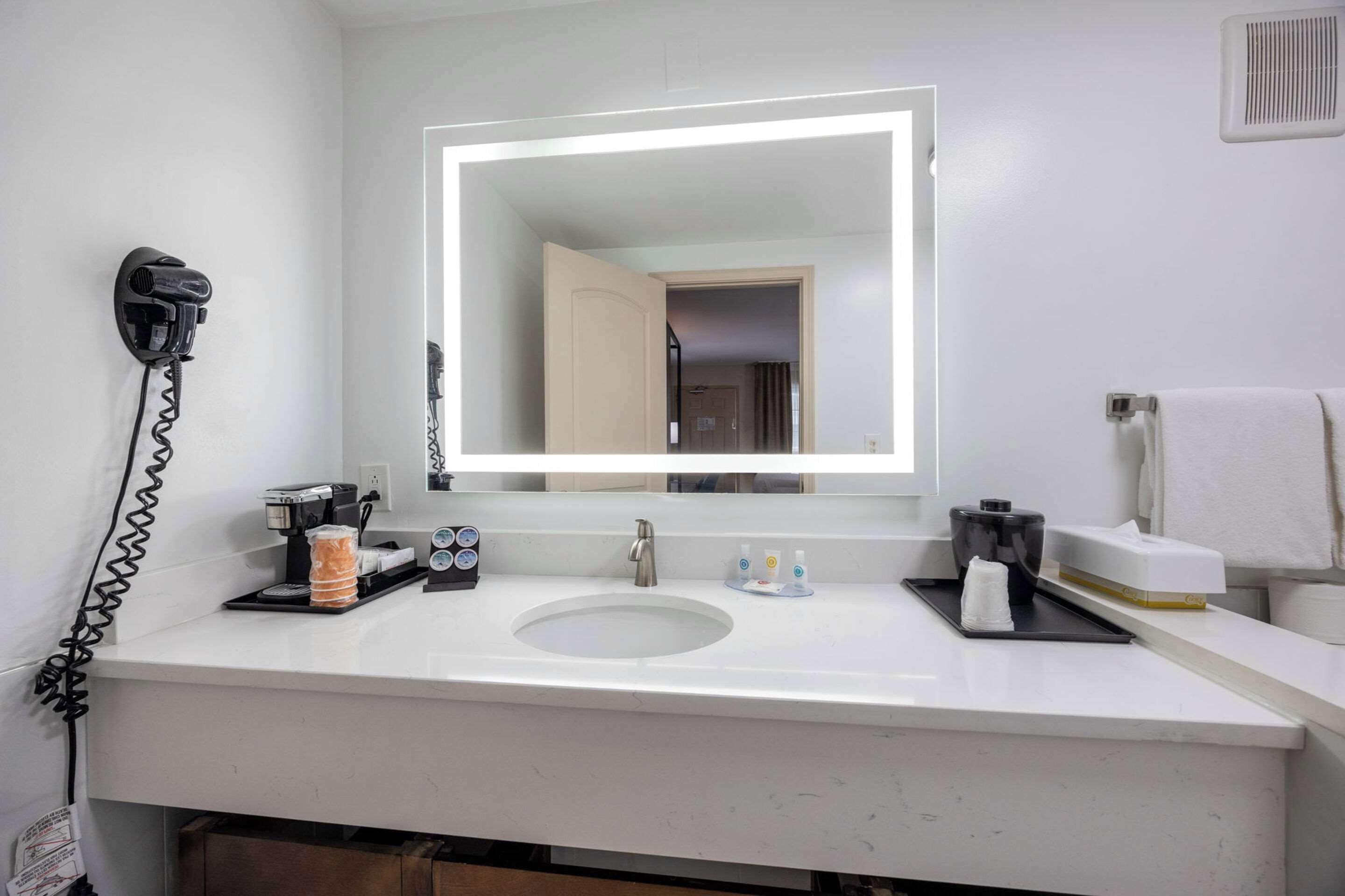 A hotel bathroom with a white sink, a large mirror with a light, a hairdryer, and toiletries on the counter.