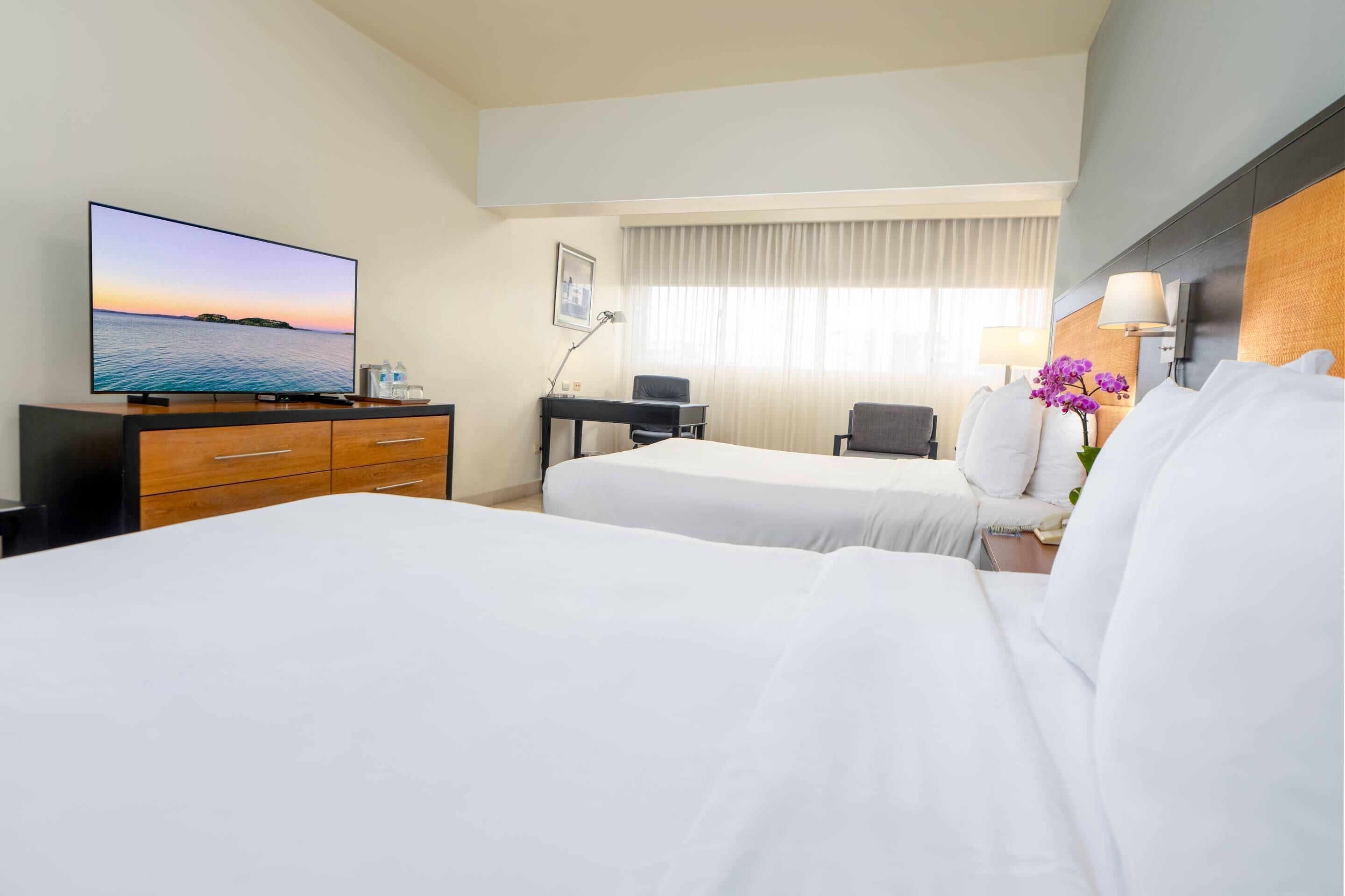 Habitación de hotel con dos camas, un escritorio, un televisor mostrando una vista del atardecer, una lámpara y una ventana con cortinas.
