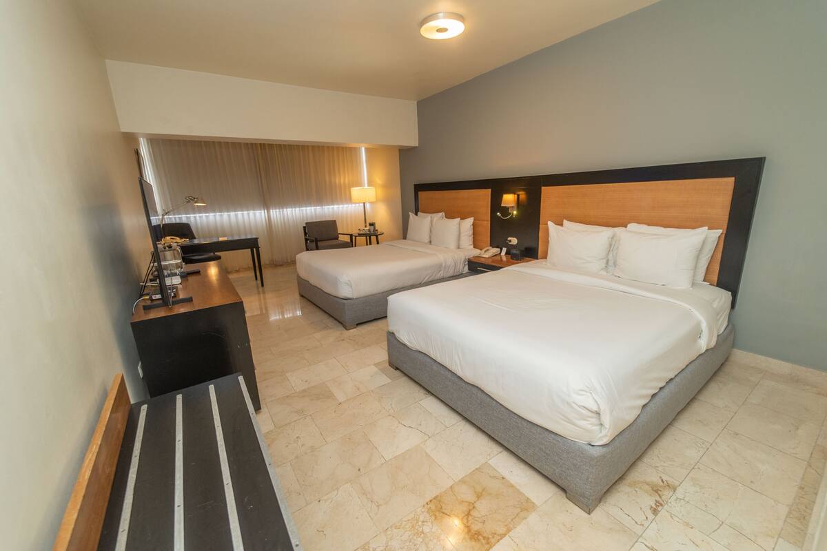 A hotel room with two beds, a desk, a chair, a lamp, and a TV.