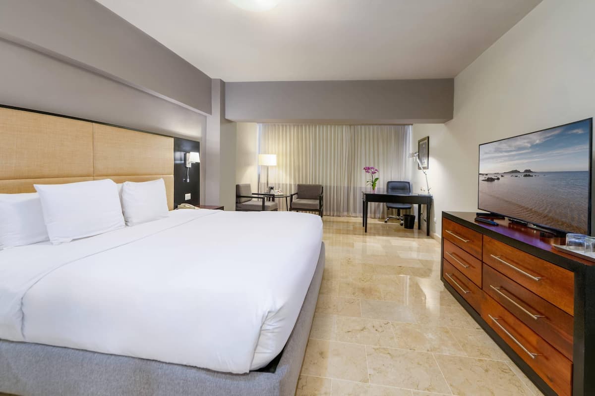 Ein Hotelzimmer mit einem großen Bett, einem Schreibtisch mit Stuhl, einem Fernseher und Meerblick.