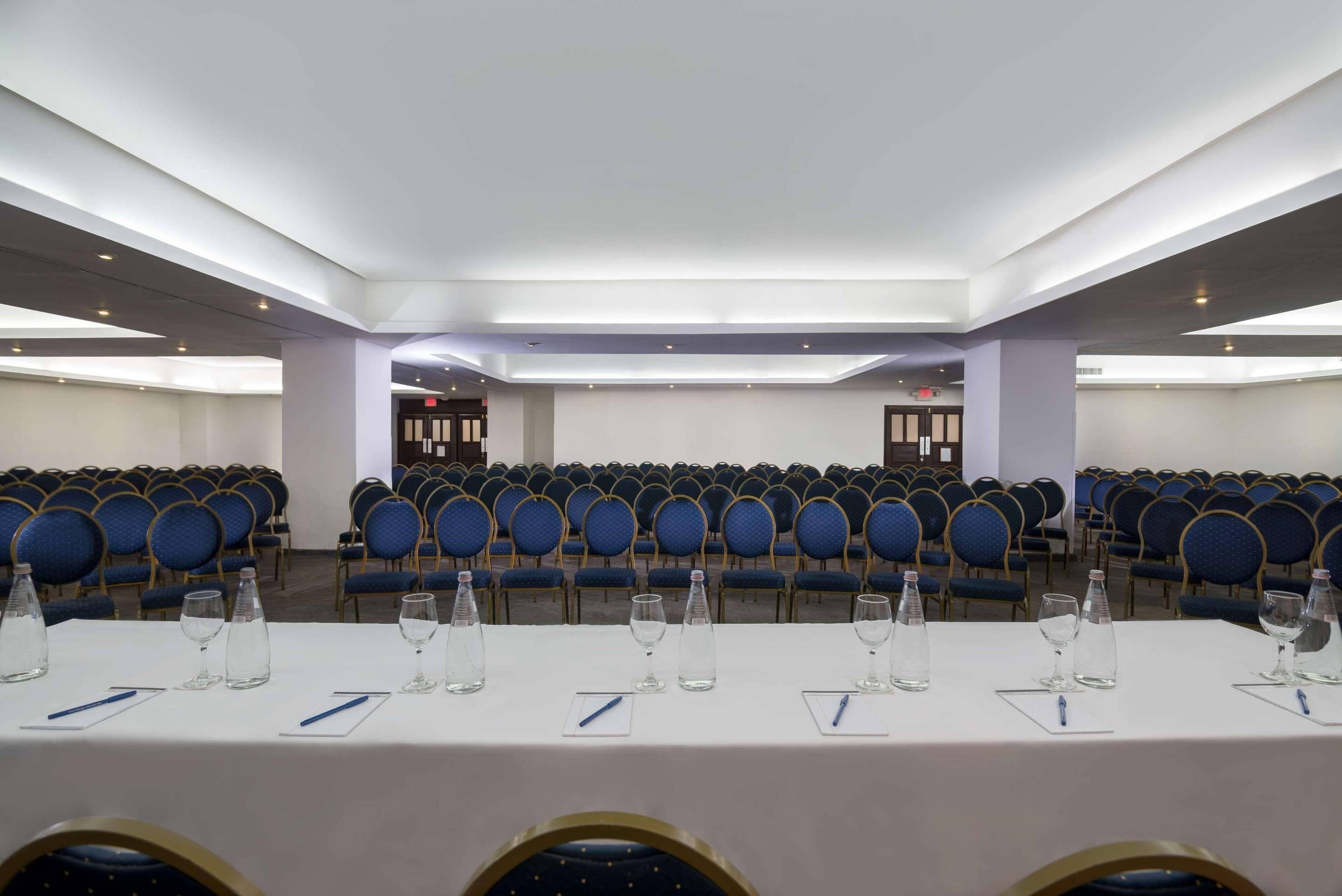 A conference room with rows of blue chairs, a long table with water bottles and pens, and a modern ceiling design.