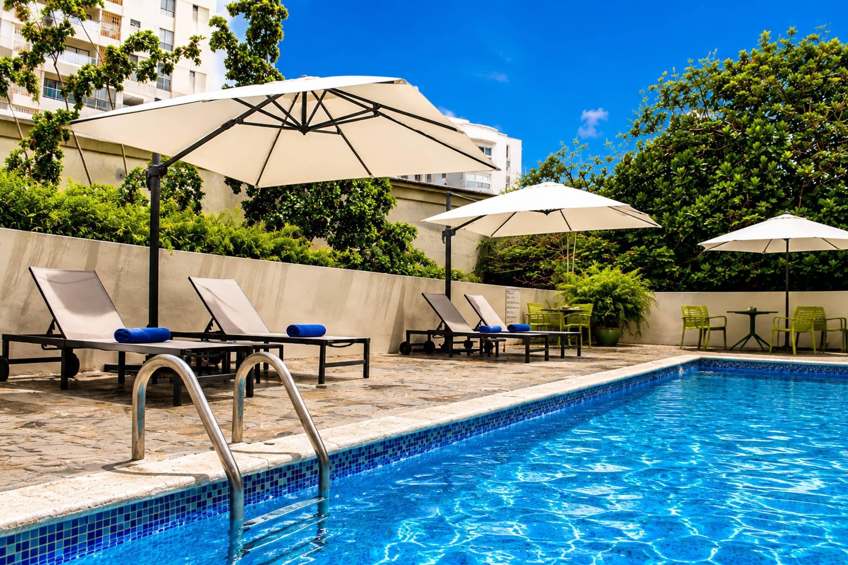 A swimming pool with lounge chairs and umbrellas, surrounded by greenery and a building.