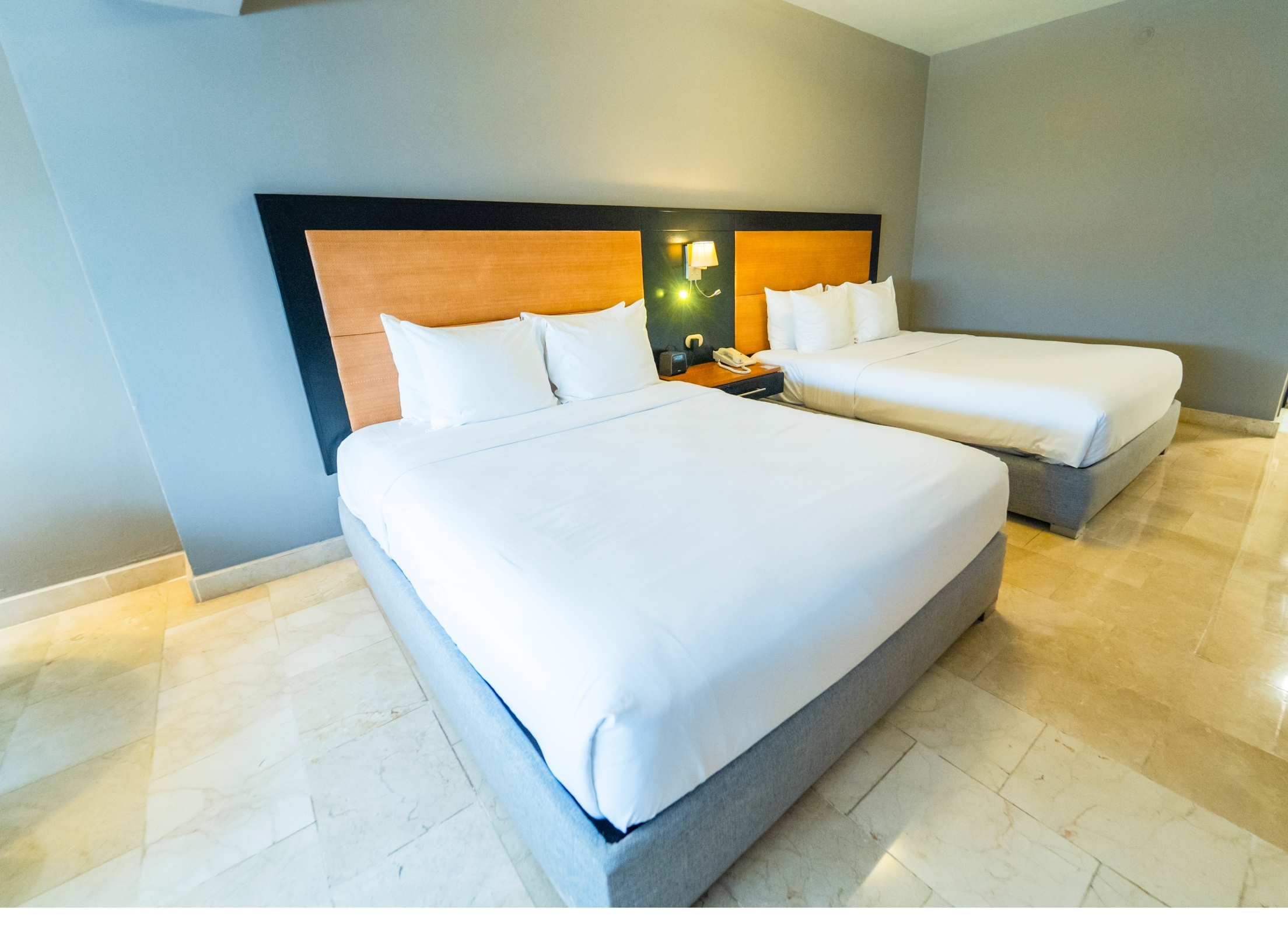 Habitación de hotel con dos camas, cabecero de madera, mesita de noche con lámpara y teléfono.