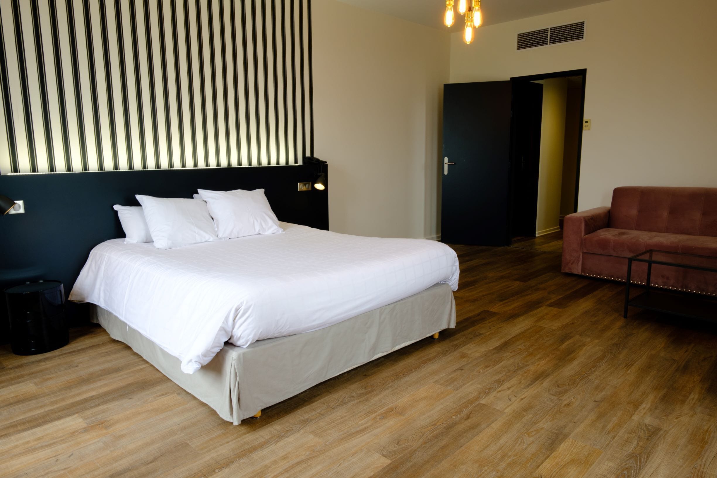 A neatly arranged bedroom with a large bed, a dark headboard, a wooden floor, a red sofa, and a door leading to another room.