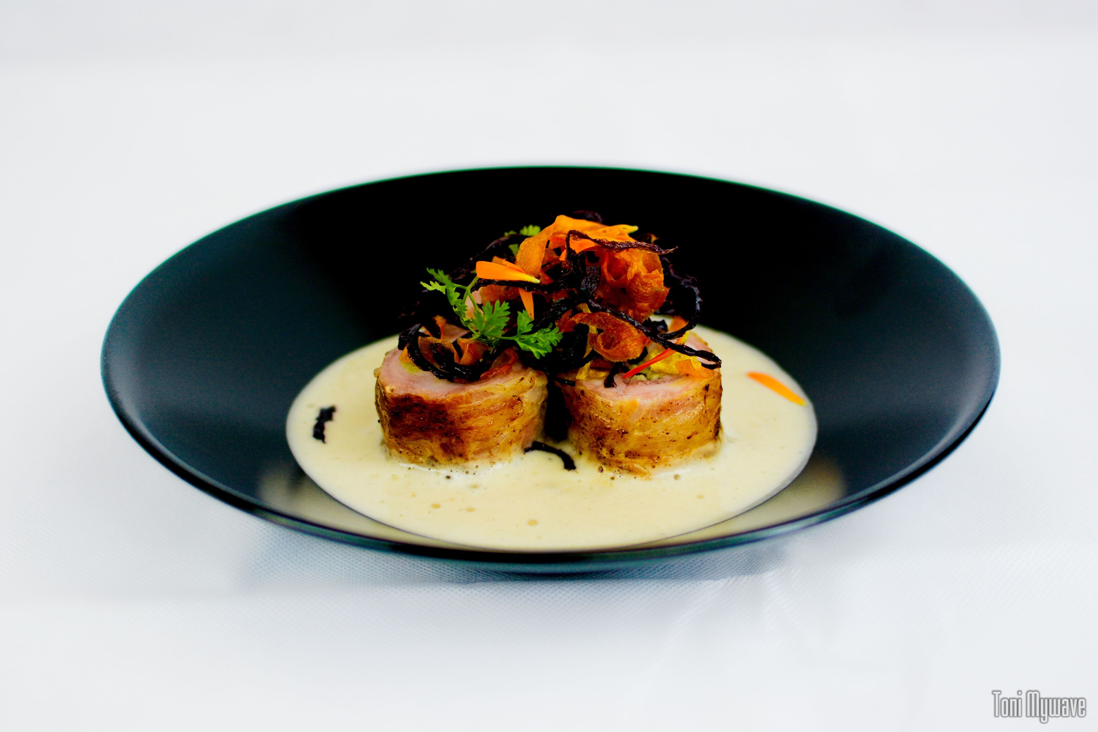 A black plate with a white sauce and two pieces of food topped with vegetables and herbs.