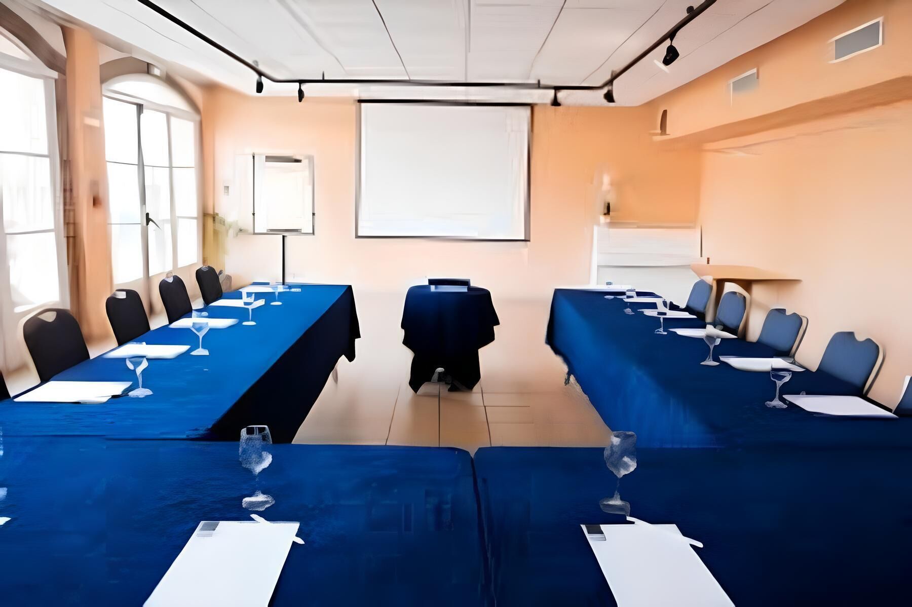 Un salón de conferencias con dos mesas largas, sillas, una pantalla de proyector y una pizarra.
