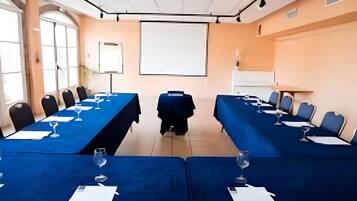 Un salón de conferencias con dos mesas largas, sillas, una pantalla de proyector y una pizarra.