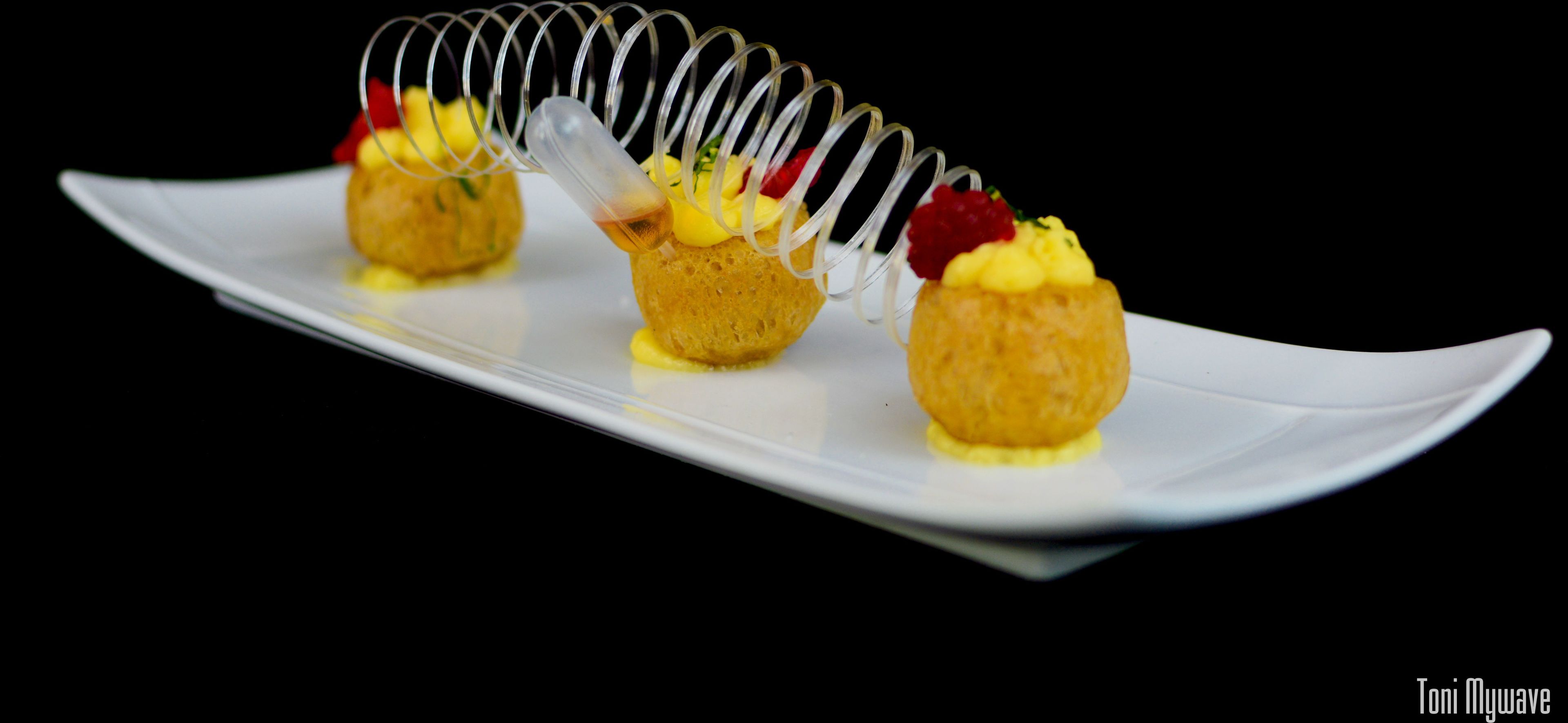 Un plato blanco con tres postres esféricos, cada uno coronado con un remolino de material transparente y decorado con elementos rojos y amarillos.