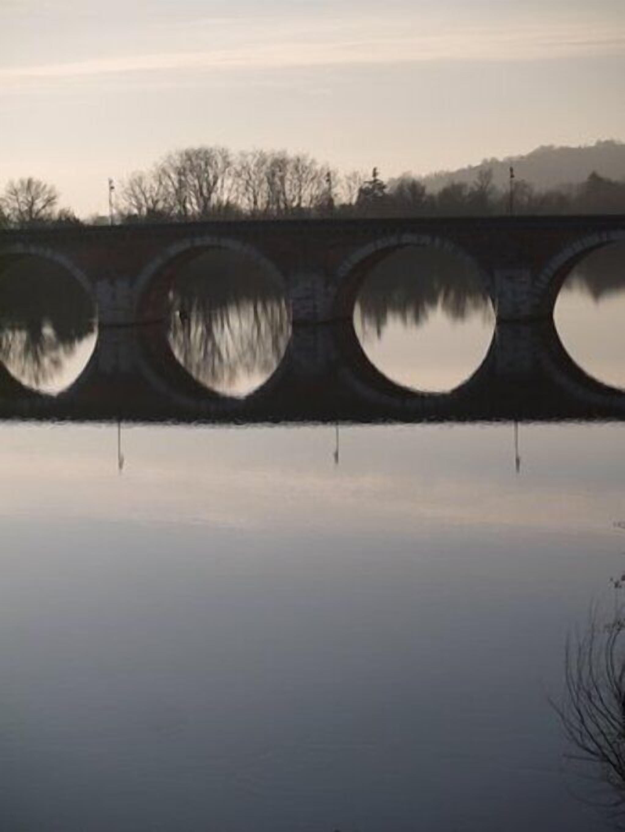 A stone bridge with circular arches over a calm body of water, reflecting the bridge and surrounding trees.