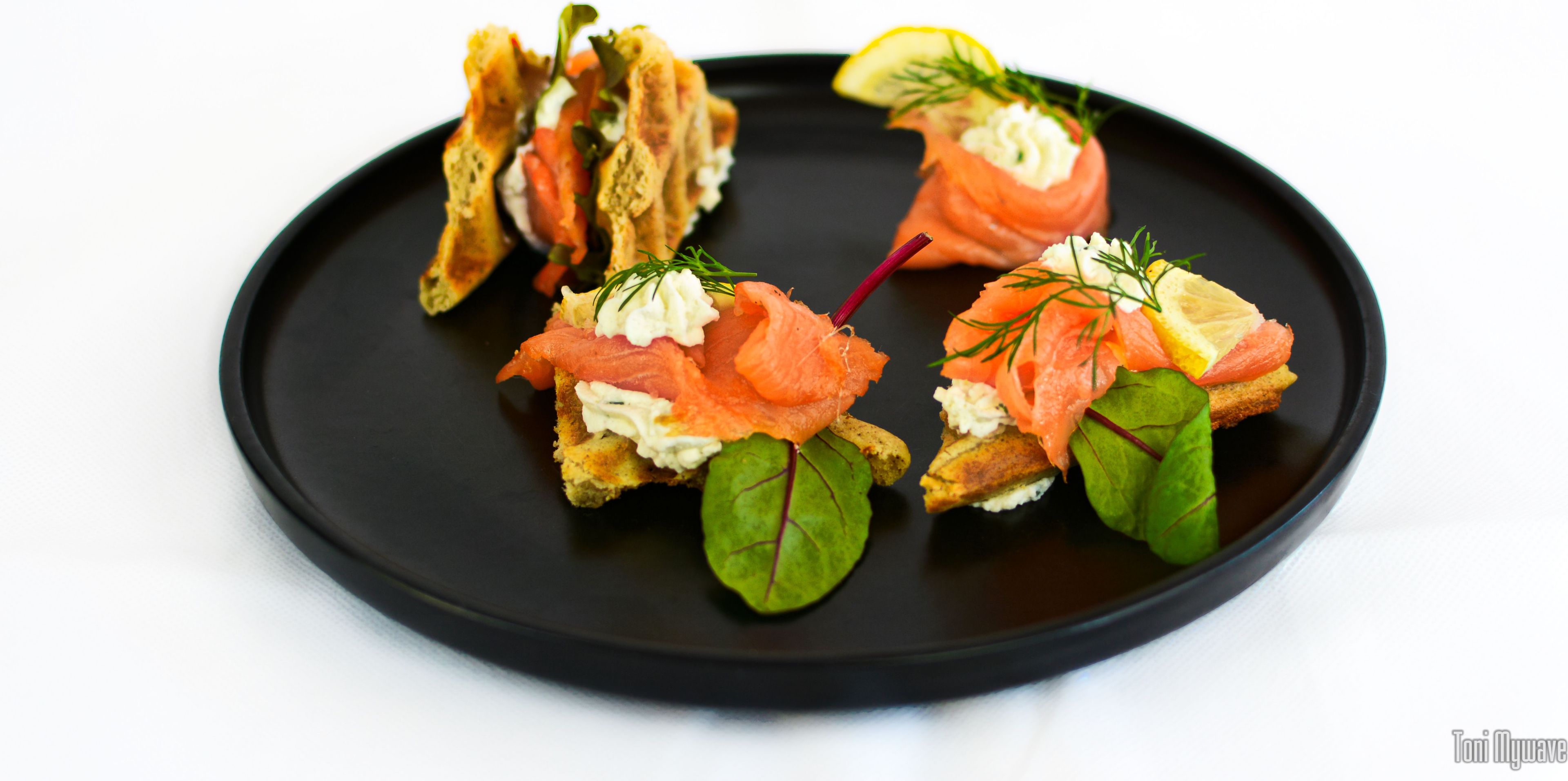 A black plate with three appetizers, each with smoked salmon, cream, and greens.