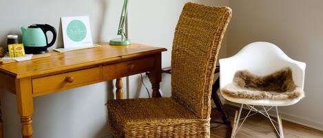 A wooden desk with a green desk lamp, a wicker chair, and a white chair with a fur throw.