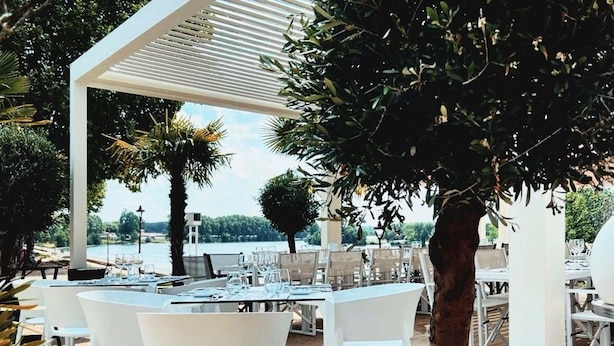 Un área de comedor al aire libre con mesas y sillas blancas, una pérgola y vista al agua.