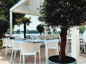 Un área de comedor al aire libre con mesas y sillas blancas, una pérgola y vista al agua.