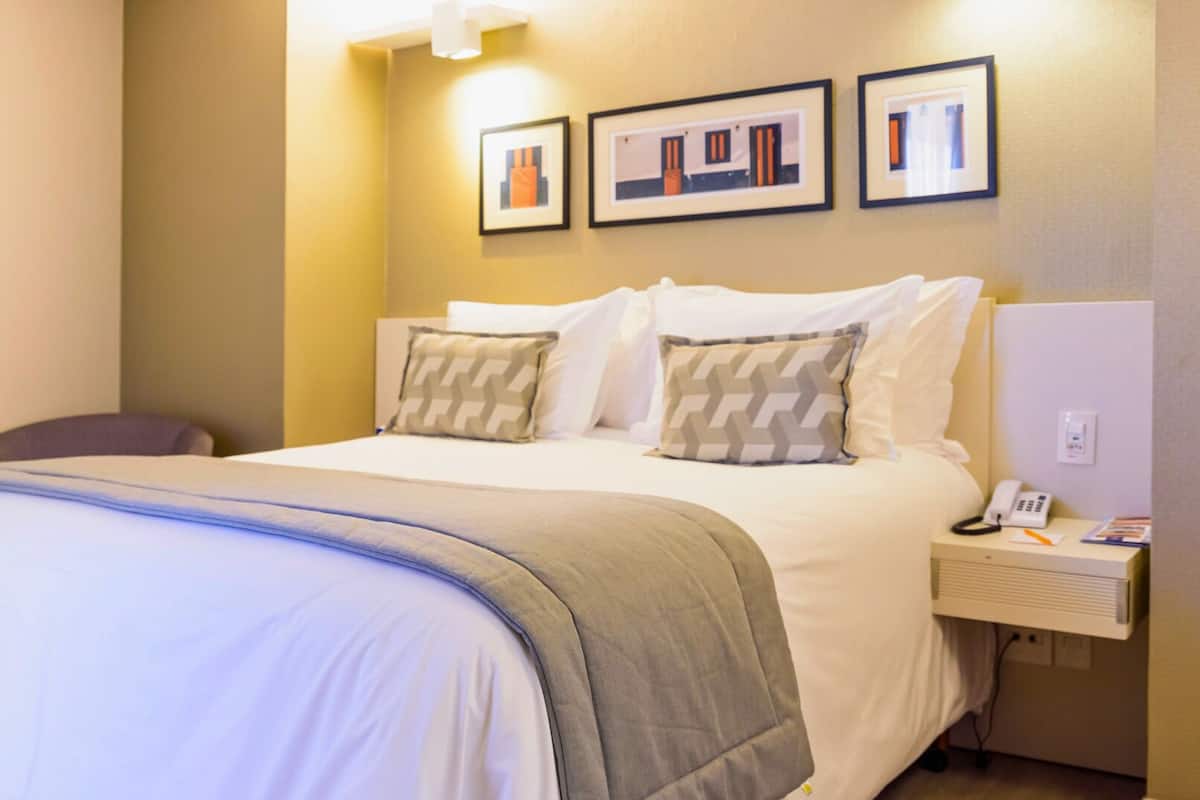Een hotelkamer met een net opgemaakt bed, een nachtkastje met een telefoon en een tijdschrift, en drie ingelijste foto's aan de muur.