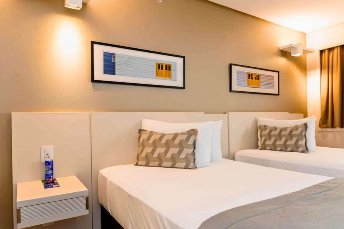Een hotelkamer met twee bedden, elk voorzien van witte lakens en gestreepte kussens. Boven de bedden hangen afbeeldingen in lijsten, en er staat een klein tafeltje met een stopcontact en een fles water.