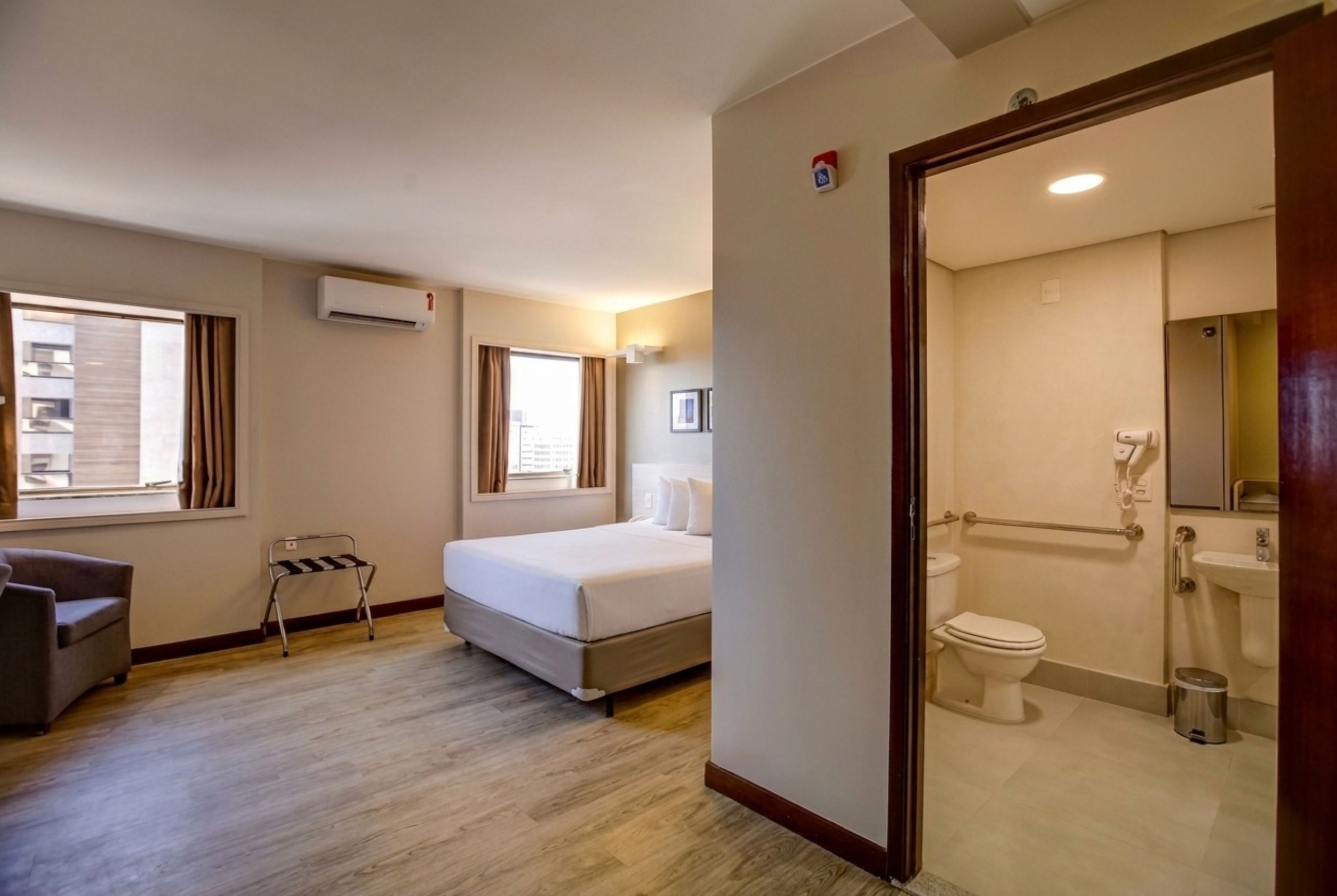 Habitación de hotel con cama, una silla, una mesita, ventana y baño con inodoro y lavabo.