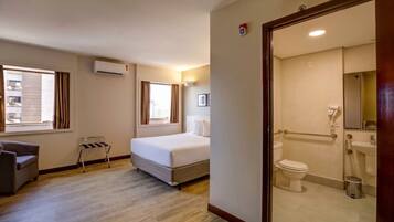 Habitación de hotel con cama, una silla, una mesita, ventana y baño con inodoro y lavabo.
